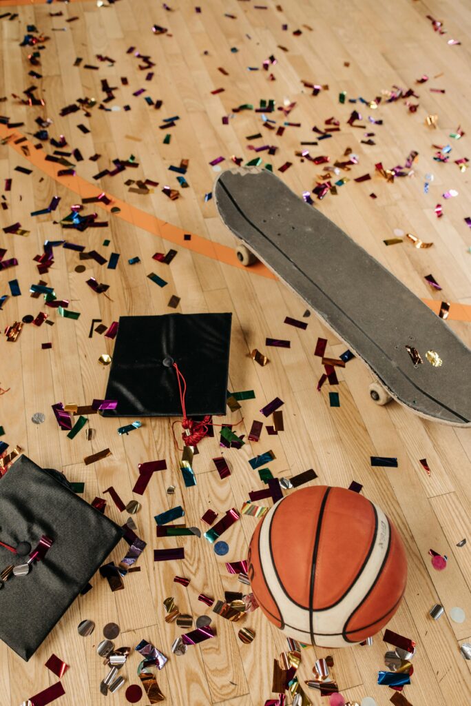 A graduation cap, basketball, and skateboard on a confetti-covered hardwood floor.